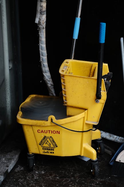 A yellow commercial cleaning mop bucket with a black removable wringer, labeled with caution symbols, positioned on a black floor outdoors next to a dark wall and a leafless tree trunk, with cleaning 