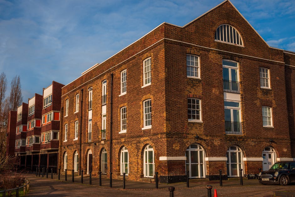 Exterior view of a multi-story brick residential building with large arched windows and balconies, illuminated by daylight against a blue sky with some clouds. The ground level features white-framed glass doors and windows, with a paved pathway and black bollards along the front. Nearby, a parked car is visible, and the area appears clean and well-maintained, reflecting professional cleaning and maintenance standards. This image exemplifies the type of residential property where Cleaners SE16 provides surface cleaning, deep cleaning, and sanitisation services, ensuring communal areas remain hygienic in line with Rotherhithe estate cleaning tips for landlords in SE16.