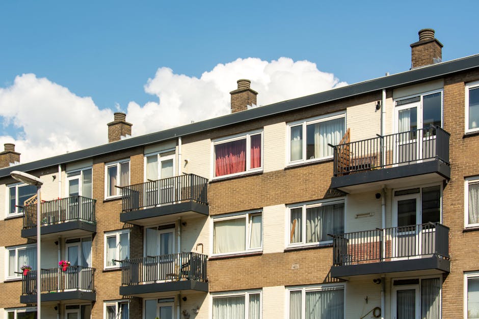 Exterior view of a multi-story brick residential building with large arched windows and balconies, illuminated by daylight against a blue sky with some clouds. The ground level features white-framed glass doors and windows, with a paved pathway and black bollards along the front. Nearby, a parked car is visible, and the area appears clean and well-maintained, reflecting professional cleaning and maintenance standards. This image exemplifies the type of residential property where Cleaners SE16 provides surface cleaning, deep cleaning, and sanitisation services, ensuring communal areas remain hygienic in line with Rotherhithe estate cleaning tips for landlords in SE16.