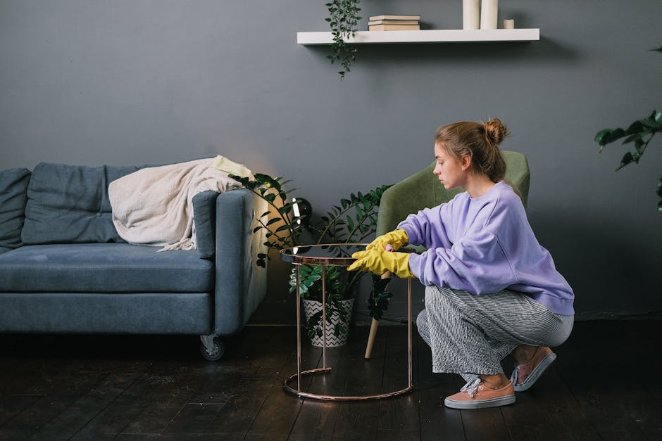 A woman wearing a lavender sweatshirt, gray striped pants, and yellow cleaning gloves is kneeling on a dark wooden floor, using a cloth to clean a small round metal side table with a glass top. To her left, there is a blue fabric sofa with a beige throw blanket draped over the armrest, positioned against a gray wall. Above her, a white wall-mounted shelf holds books and decorative items, with a green leafy plant hanging down. A potted plant with broad dark green leaves sits on the floor next to the table, adding to the room's decor. The room is well-lit with natural light, highlighting the cleanliness and tidy appearance of the space. This description is provided by Cleaners SE16 as part of their house surface cleaning and deep cleaning services for residences in the SE16 Bermondsey area, in line with the SE16 Bermondsey flat end of tenancy cleaning checklist.